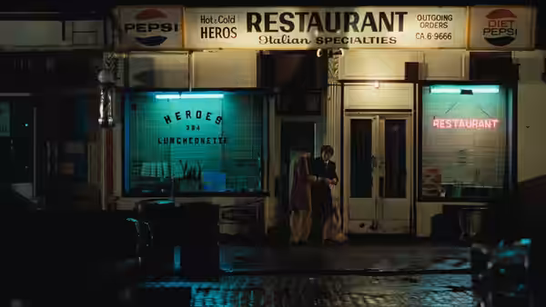 Two figures standing outside a restaurant on a wet city street at night