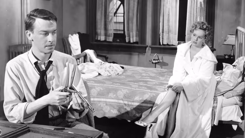 Bart Tare (John Dall) examines a revolver as Annie Laurie Starr (Peggy Cummins) sits on a bed