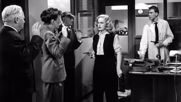A woman faces several men in an office while a man stands behind the counter