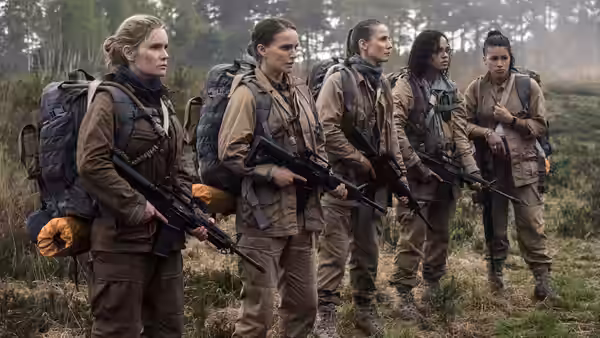 Five women in fatigues with rifles and backpacks stand in formation outdoors