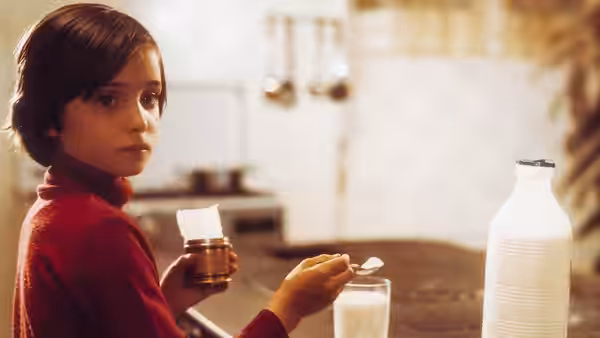Ana (Ana Torrent) sits at a kitchen table with a glass of milk and a spoon