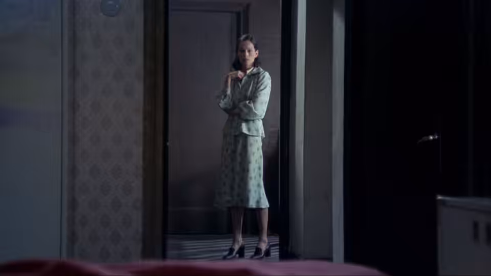 María (Geraldine Chaplin) stands in a doorway, clutching her hands in a dim room
