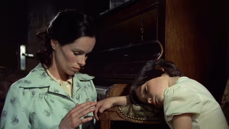 María (Geraldine Chaplin) examines a child's hand beside a piano while the child rests her head