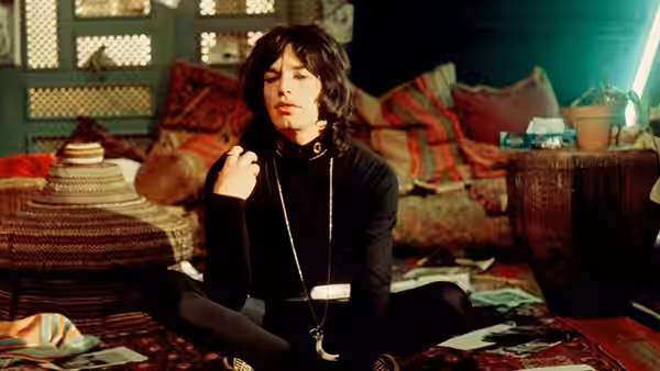 Turner (Mick Jagger) sits cross-legged on a rug adjusting his necklace