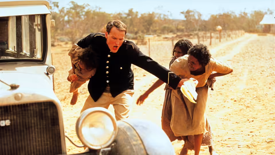 A uniformed man reaches for two girls beside a parked car on a dusty road