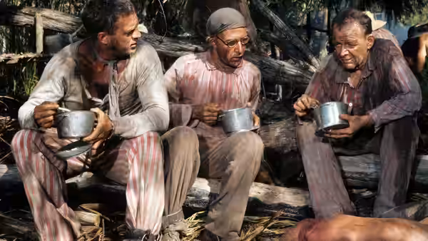 Papillon (Steve McQueen) and two prisoners eating from metal bowls outdoors