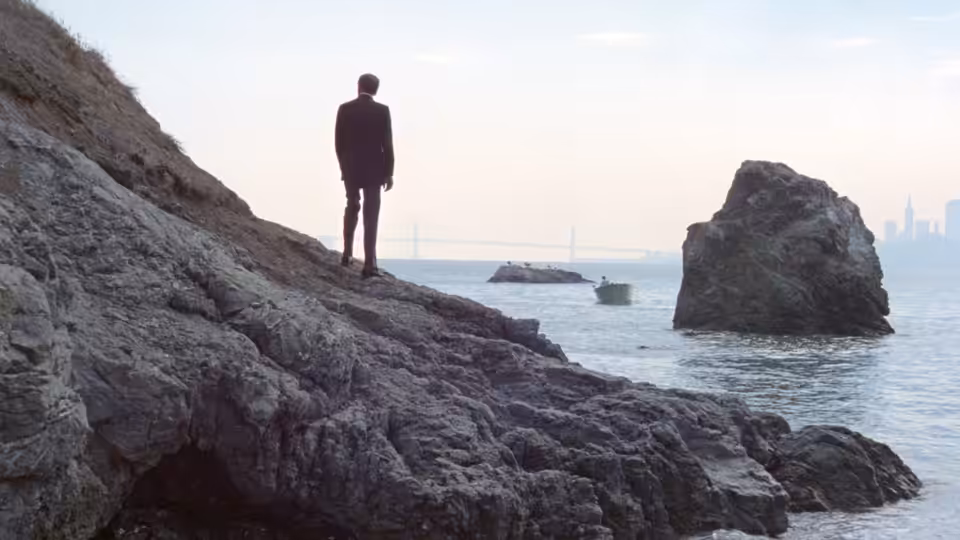 Frank Morris (Clint Eastwood) stands on rocky shore, facing the water beyond Alcatraz