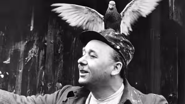 A smiling man in a cap stands beneath a pigeon spreading its wings