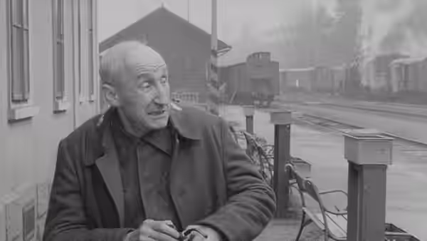 An older man in a coat stands by railway tracks and looks to the side