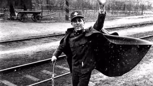 A uniformed railway worker waves beside the tracks, holding an umbrella open overhead