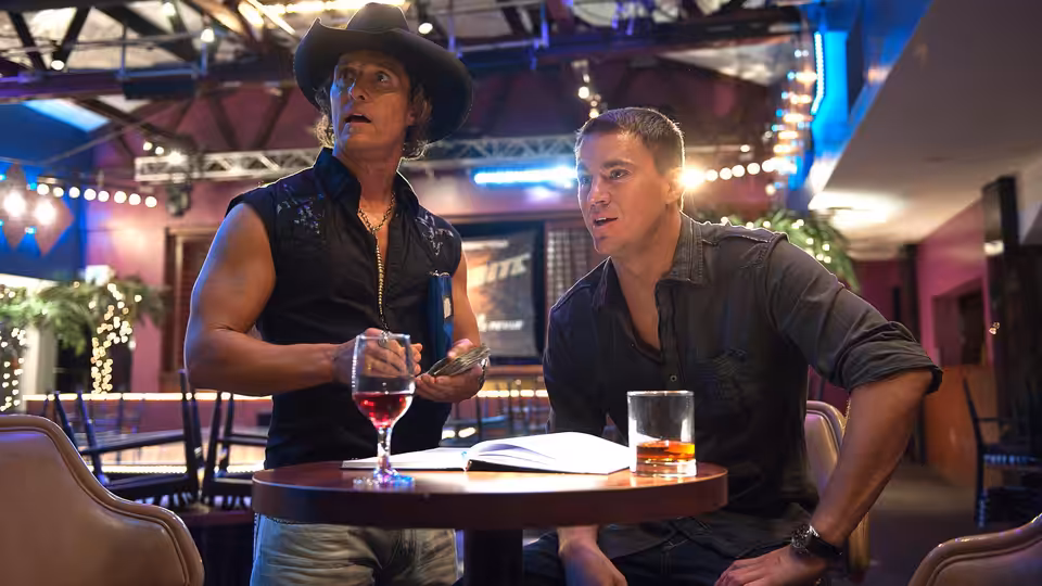 Dallas (Matthew McConaughey) and Mike (Channing Tatum) seated at a table with drinks