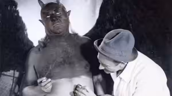 A bare-chested man in a horned mask stands beside a man in a white hat holding a pencil