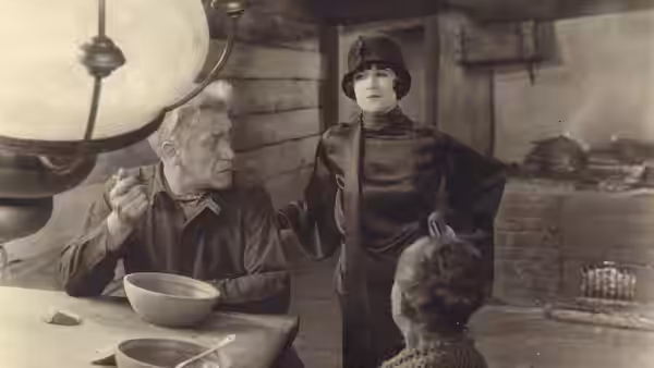 An older man eats from a bowl at a table as a woman stands nearby