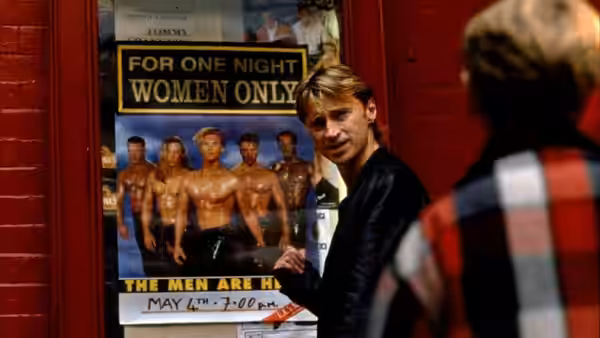 Guy (Hugo Speer) turns toward a person in a plaid shirt near a poster