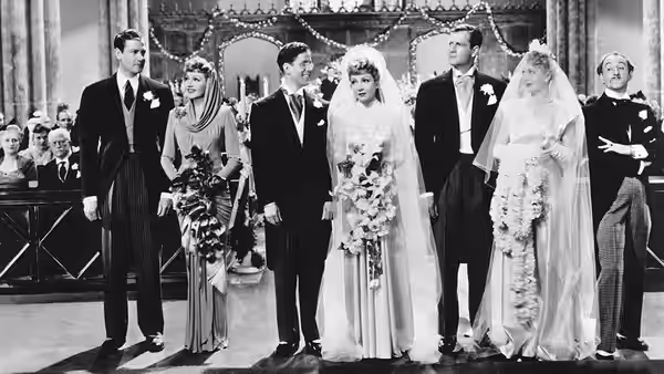 A wedding party stands in a row beneath a decorated arch, with two brides and their attendants