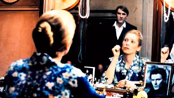 A woman studies her reflection at a dressing-room vanity as a man stands behind her