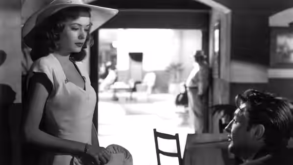 A woman in a white dress and hat faces a seated man with a cigarette