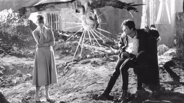 A woman stands among rubble while a young man sits in a chair nearby