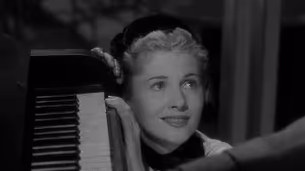 Lisa Berndle (Joan Fontaine) leans against a piano with one hand on the keys