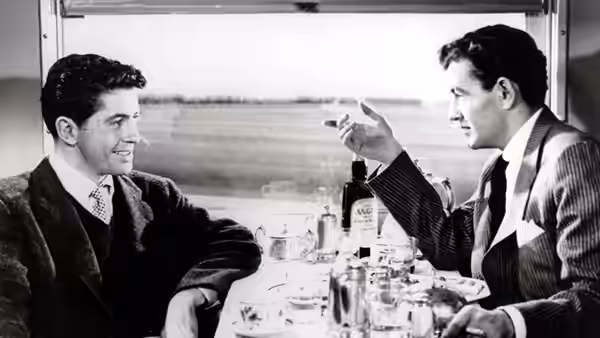 Two men talk over drinks at a table aboard a moving train