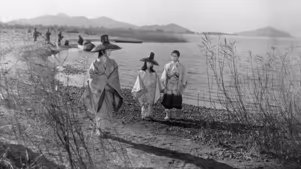 Three women in traditional hats and robes walking along a lakeside path