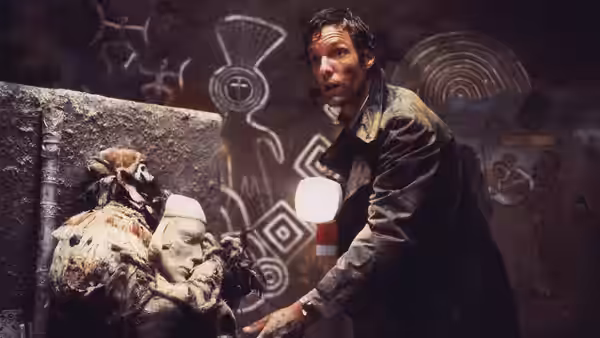 David Burton (Richard Chamberlain) examines carved figures and symbols in a dim cave