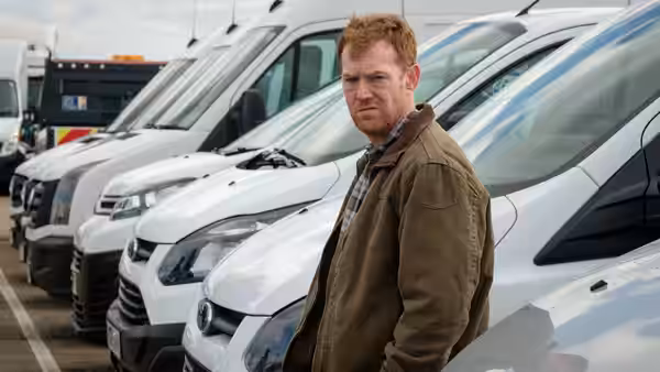 Ricky (Kris Hitchen) stands among a row of white delivery vans