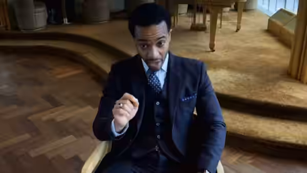 Ray Burke (André Holland) gesturing from a chair in a spacious lobby