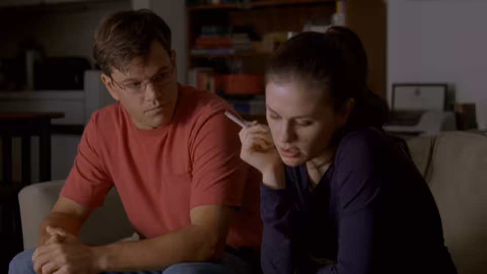 Man in red shirt and woman in blue top sit on a couch as she holds a phone