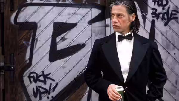 Cahit Tomruk (Birol Ünel) in a tuxedo holds a drink before a graffiti-covered door