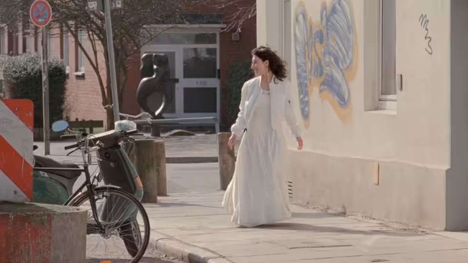 Sibel Güner (Sibel Kekilli) in a white dress walks past a mural and parked bicycle