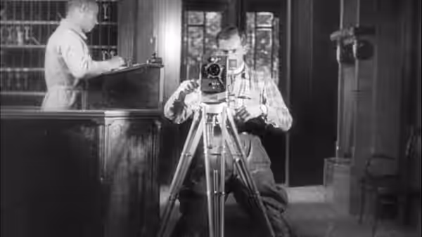 The Cameraman (Mikhail Kaufman) adjusts a tripod-mounted camera inside a room
