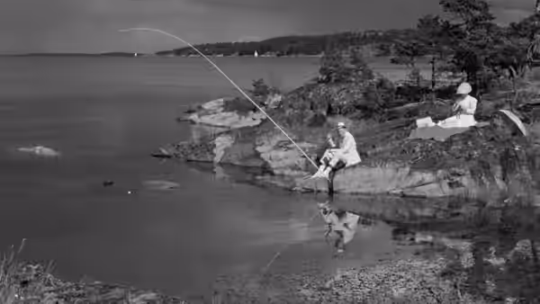 A man fishes from the rocky shore while another person sits beside the water