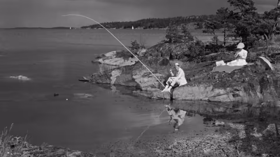 A man fishes from the rocky shore while another person sits beside the water