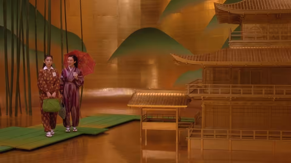 two figures in traditional robes stand on green mats before a golden screen and pagoda model