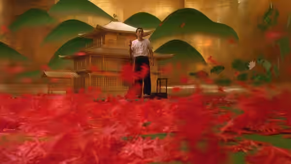 man stands amid floating red petals in a golden room with a temple model behind him