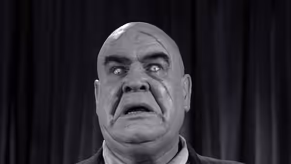 A bald man with wide eyes stares forward with open mouth