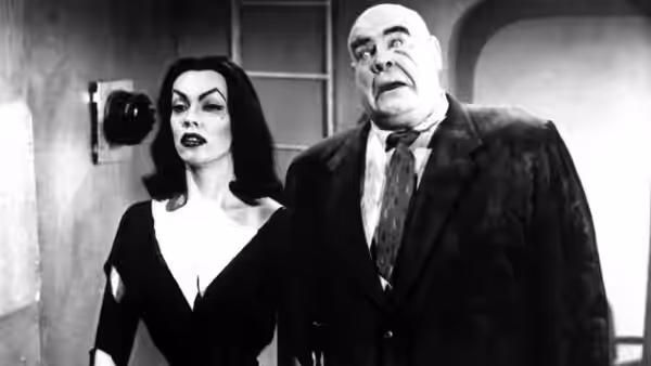 A woman in a black dress stands beside a bald man in a suit