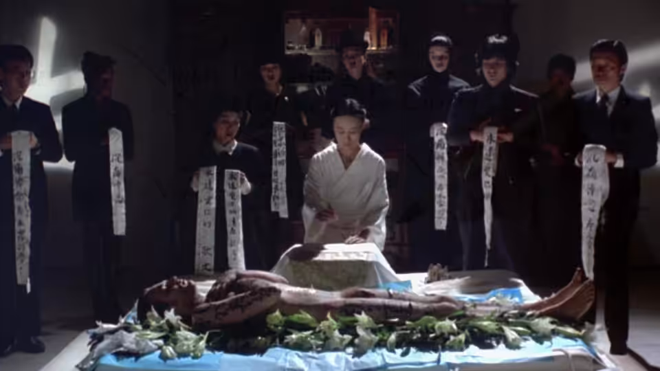 A man in white kneels beside a body as suited men hold strips of calligraphy