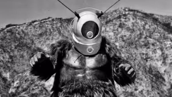 Ro-Man stands with arms raised against rocky hills under clear sky
