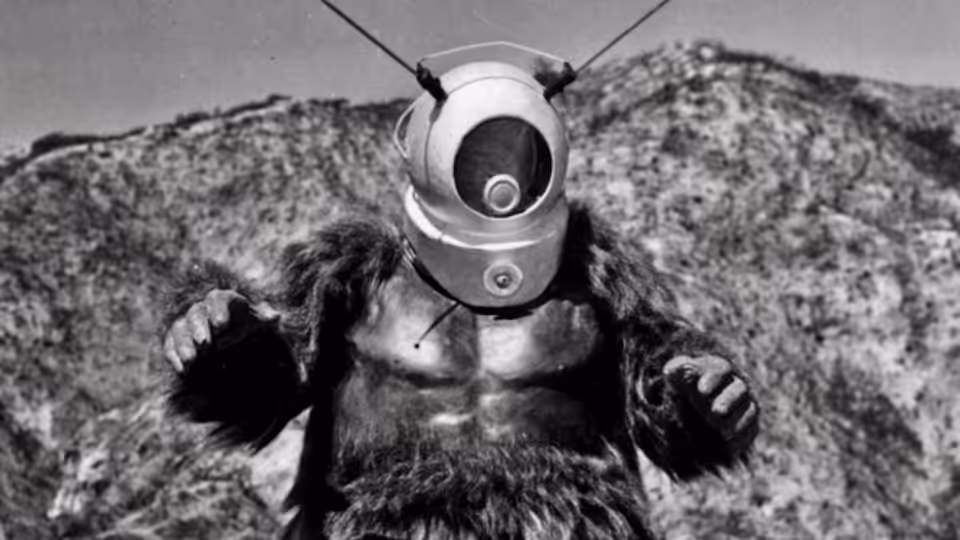 Ro-Man stands with arms raised against rocky hills under clear sky