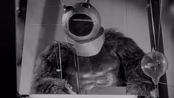 Ro-Man seated at table with wires and glass equipment indoors