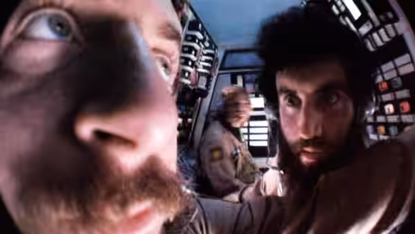 Two bearded crewmen peer toward the camera inside a spacecraft control room