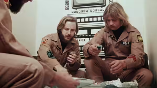 Two crewmen in tan uniforms examine a small object on the spacecraft floor