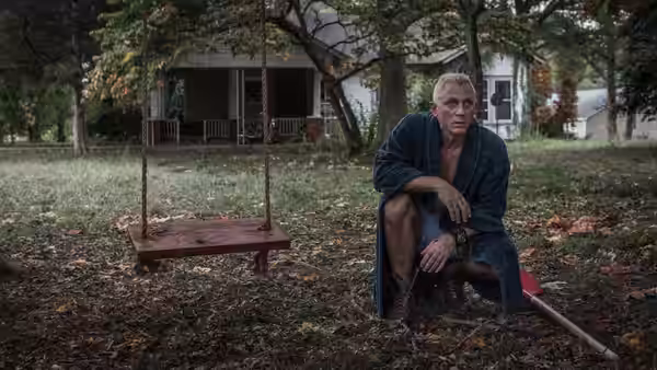 Joe Bang (Daniel Craig) crouching in a yard beside a swing set