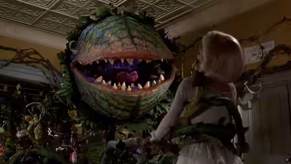 A blonde woman facing a large plant with sharp teeth and open mouth