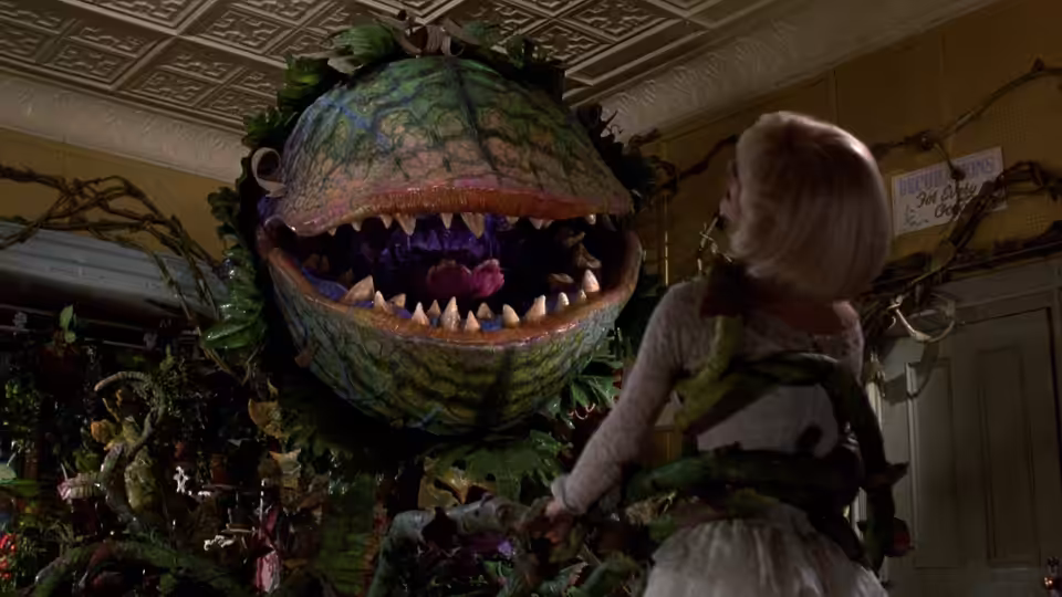 A blonde woman facing a large plant with sharp teeth and open mouth
