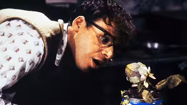 Seymour Krelborn (Rick Moranis) leaning over a wilting plant in a can