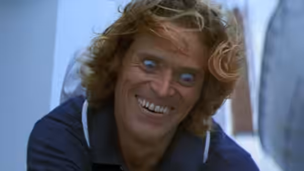 Geiger (Willem Dafoe) grinning with wild blond hair and bright eyes