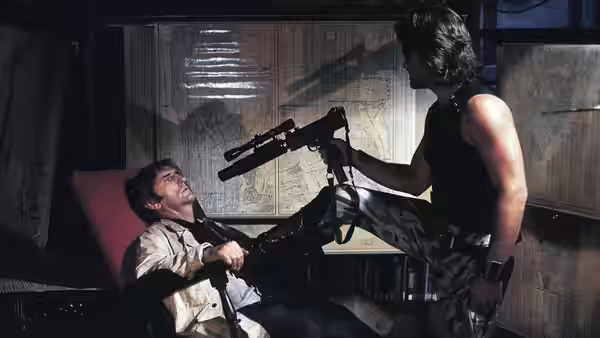 Snake Plissken (Kurt Russell) points a rifle at Brain (Harry Dean Stanton), seated beneath a map